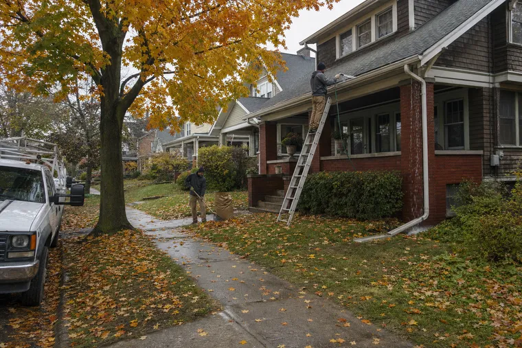 Technician flushing a bungalow's porch gutter with a hose while a second bags leaves on the lawn